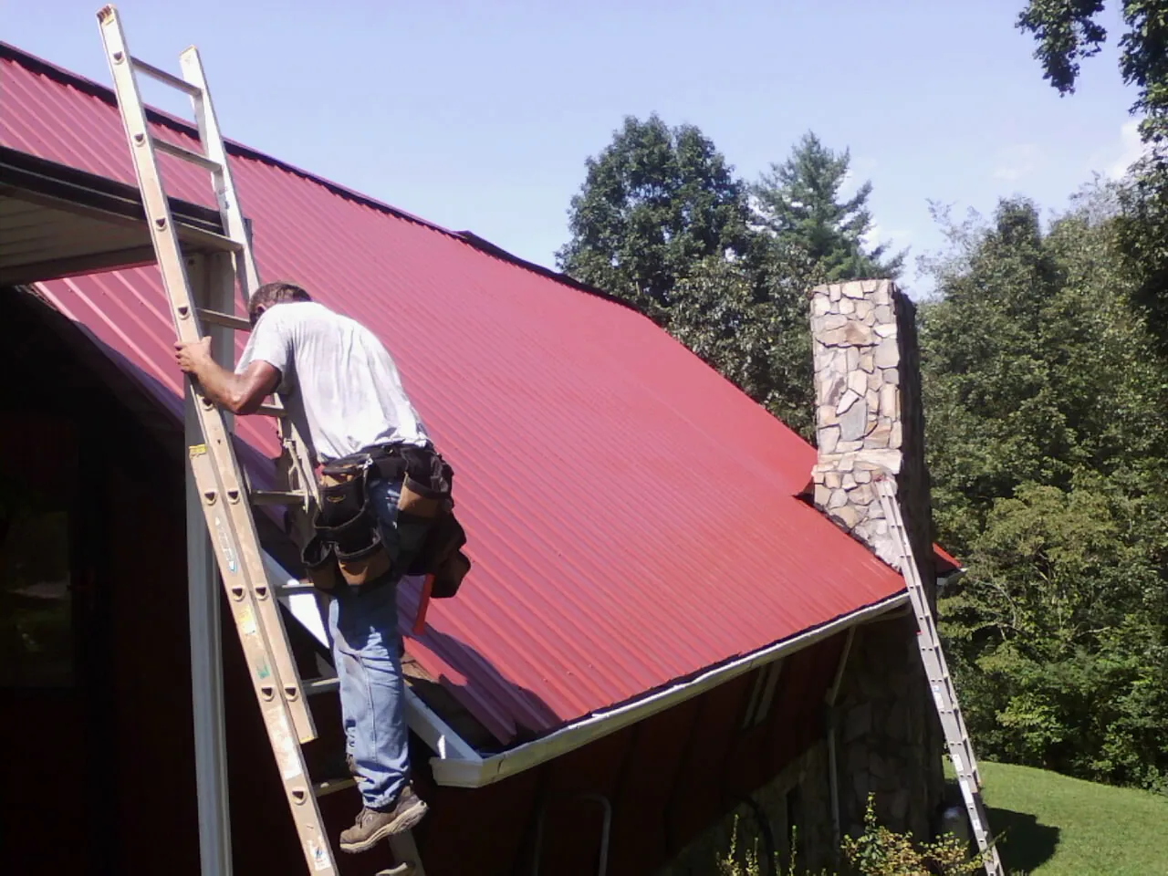 Roofer on a ladder beside a red metal roof and stone chimney — Soffit & Fascia Repair by Roofing Brothers in North Olmsted